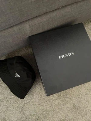Svart bucket hat från Prada - Säljer en svart bucket hat från Prada i glansigt nylonmaterial. Klassisk form med brett brätte och ikoniska metalltriangeln med logga framtill. Perfekt accessoar för en trendig och stilren look.