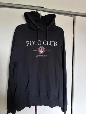 Svart hoodie från Polo Club Saint Vincent - Snygg svart hoodie från Polo Club Saint Vincent med tryck på bröstet där det står 'POLO CLUB' och 'SAINT VINCENT'. 