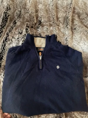 Marinblå halvzip tröja från Morris - Snygg marinblå stickad tröja från Morris med halvzip och beige insida i kragen. Tröjan har en liten broderad logga på bröstet och ribbade muddar. Perfekt för en stilren och avslappnad look.