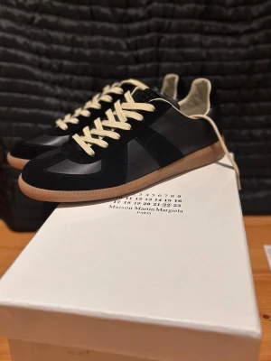 Maison Margiela gats - Stilrena svarta sneakers från Maison Margiela med beigea skosnören och gummisula. Ovandelen är i en mix av mocka och läder, vilket ger en lyxig känsla. Klassisk siluett med låg profil och diskreta detaljer. Perfekta för dig som gillar minimalistisk design.