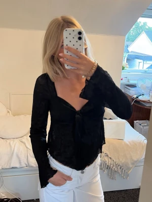 Svart långärmad topp med knyt - Trendig svart långärmad topp med djup urringning och knyt framtill. Figurnära passform och lätt transparent material som ger en cool vibe. Perfekt att styla med jeans eller kjol för en snygg look.