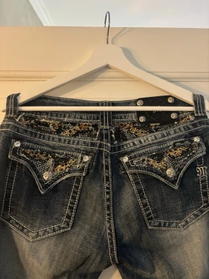 Blå jeans med stenar - Säljer ett par blå jeans med leopardmönstrade detaljer och glittriga stenar på bakfickorna. Några stenar har lossnat genom åren. Jeansen har kontrastsömmar och bootcut-modell som ger en snygg siluett. Perfekta för dig som vill sticka ut lite extra! Innerbenslängd: 84cm, midjemått 38cm