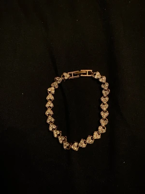 Guldarmband med hjärtformade stenar - Snyggt armband i guldfärg med hjärtformade länkar, där varje hjärta är dekorerat med glittrande stenar. Perfekt accessoar för dig som gillar detaljer och vill ha något som sticker ut. Låset är diskret och smidigt. Passar till många olika stilar.