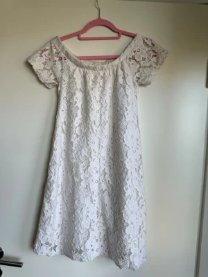 Vit spetsklänning med kort ärm från River  Island - Romantisk vit klänning i spets med blommigt mönster och offshoulder-ringning. Klänningen har korta ärmar och en rak, luftig passform som slutar vid knät. Perfekt för sommarens alla tillfällen och ger en fräsch och trendig look.