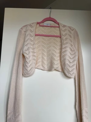 Ljusbeige kort bolero i spetsstickning - Supersöt ljusbeige bolero med spetsstickat mönster och lång ärm. Kort och öppen framtill, perfekt att slänga över en klänning eller topp. Materialet är tunt och luftigt, vilket ger en fräsch och trendig look.