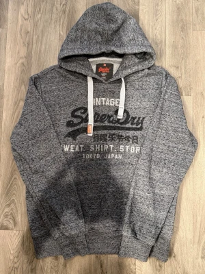 Superdry Vintage Hoodie Mörkgrå Strl XL (Passar L/XL) - Superdry Vintage mörkgrå hoodie med klassisk passform, märkt storlek XL men passar bäst som rymlig L eller normal XL. Axelbredd 52 cm, bröstvidd 60 cm, armlängd 69,5 cm, midja 47,5 cm, längd 72 cm, i fint skick.