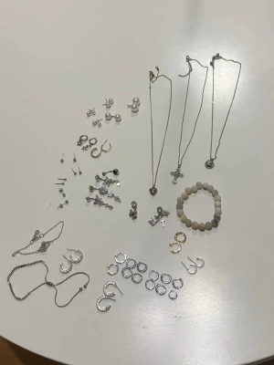 Mixat smyckespaket i silver  - Stort paket med olika smycken: halsband med hjärta, kors och medaljong, flera par örhängen i olika former (ringar, stift, pärlor), armband i silverfärg och ett pärlarmband i vitt och beige.