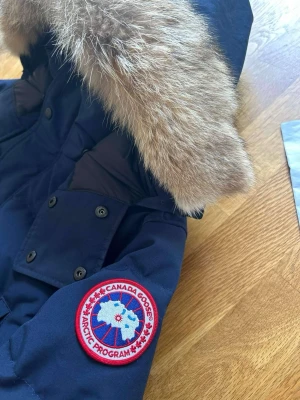 Mörkblå Canada Goose wyndham parka med päls - Snygg mörkblå wyndham parka från Canada Goose med stor luva och äkta pälsdetalj. Jackan har flera fickor med knappar och den klassiska Canada Goose-loggan på ärmen. Perfekt för kalla vinterdagar och riktigt varm.