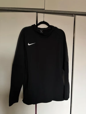 Svart långärmad Nike Dri-FIT tröja - Säljer en svart långärmad träningströja från Nike med Dri-FIT material som transporterar bort fukt. Tröjan har en enkel design med vit Nike-logga på bröstet och rund hals. Perfekt för träning eller sportaktiviteter.