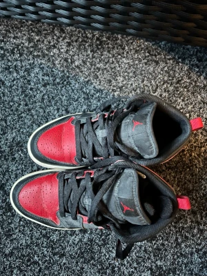 Svart-röda Air Jordan sneakers - Snygga Air Jordan sneakers i svart och rött läder med klassisk hög siluett. Skorna har svarta skosnören, vit sula och den ikoniska Jumpman-loggan på plösen. Perfekta för dig som vill ha en streetwear-look med retrovibbar.