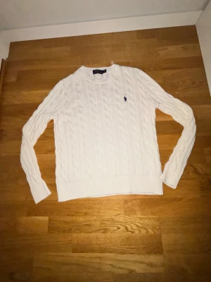 Vit kabelstickad tröja Polo Ralph Lauren - Kabelstickad vit tröja från Polo Ralph Lauren med rund halsringning och lång ärm. Tröjan har det klassiska broderade logotypen på bröstet och ribbade muddar vid ärmslut och nederkant. Perfekt för en clean och stilren look.