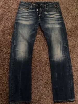 Blå Replay jeans med slitningar - Jeansen är slitna och har ett hål vid skrevet, slitna vid nedre vaderna som går att laga därav priset. Första bilderna är några månader gamla. Riktigt fet passform som sitter redigt bootcut. 