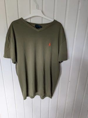 Olivgrön v-ringad t-shirt från Ralph Lauren  - Olivgrön t-shirt med v-ringning och korta ärmar. Tillverkad i 100% bomull och har en liten broderad logga i orange på bröstet. Perfekt för en avslappnad och stilren look.