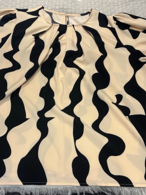 Beige blus med svart vågmönster - Säljer en beige blus med svart vågigt mönster och rund halsringning. Blusen har långa ärmar med resår vid ärmslut och ett luftigt, avslappnat fall. Perfekt för dig som gillar statement-plagg och grafiska prints.