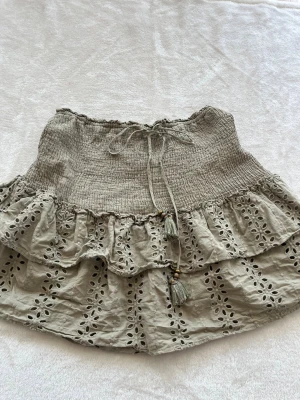 Grön kortkjol med volanger och broderi - Supersöt grön kortkjol med volanger och broderade blommönster. Kjolen har smock i midjan och dekorativ knytning med tofsar. Perfekt för sommarens alla tillfällen och ger en bohemisk vibe.