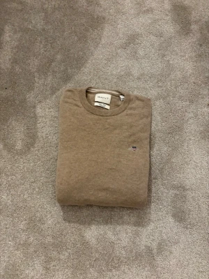 Beige stickad tröja från Gant - Klassisk beige stickad tröja från Gant med rund halsringning och liten broderad logga på bröstet. Tröjan har långa ärmar och ribbade muddar vid ärmslut och nederkant. Perfekt för lager på lager och enkel att matcha.