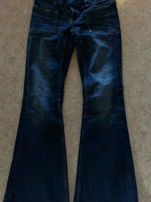 Mörkblå bootcut jeans med fickor - Snygga mörkblå bootcut jeans med två stora framfickor och dubbla knappar i midjan. Jeansen har en lätt tvättad look och utsvängda ben, perfekta för en trendig och avslappnad stil.