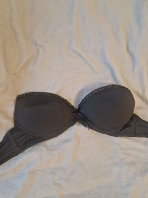 Svart axelbandslös push up bh - Säljer en svart axelbandslös bh från Lormar med lätt vadderade kupor och spetskant upptill. Bh:n har en liten rosett framtill och breda sidopartier för extra stöd. Perfekt under toppar utan axelband.