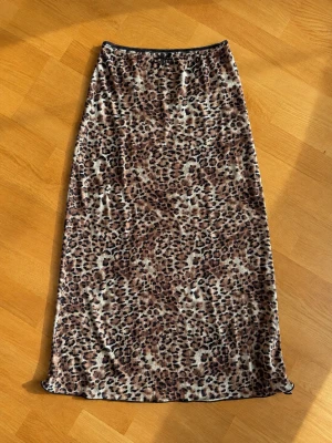 Leopardmönstrad långkjol med rosett - Snygg långkjol med leopardmönster i bruna och svarta toner. Kjolen har en liten svart rosett i midjan och resår för skön passform. Perfekt för dig som vill sticka ut med ett djurmönster. Materialet känns mjukt och följsamt.