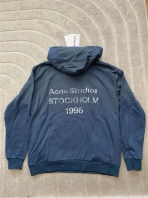Blå hoodie från Acne Studios - Snygg blå hoodie från Acne Studios med tryck på ryggen där det står 'Acne Studios STOCKHOLM 1996'. Hoodien har huva, långa ärmar och ribbade muddar. Tillverkad i mjukt bomullsmaterial och har en clean look utan fickor framtill. Köppt på plick men ganska säker på att den är äkta!
