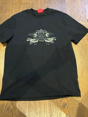 Svart t-shirt med tryck från Hugo - Svart t-shirt från Hugo med ett detaljerat tryck framtill som föreställer två fiskar och blommor. Klassisk passform med rund hals och korta ärmar. Tillverkad i mjuk bomull för en bekväm känsla. Storlek xs men passar perfekt på dig som burkar ha s! Har användts 2 gånger 