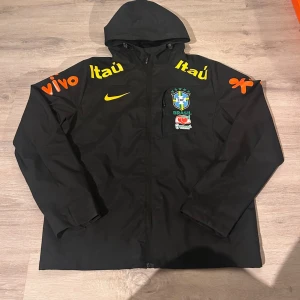 Svart Brasil Nike vindjacka med huva - Snygg svart vindjacka från Nike med huva och dragkedja. Jackan har Brasilien CBF-logga, gula och orangea sponsortryck samt en ficka på bröstet. Perfekt för dig som gillar sportig stil och vill sticka ut med färgglada detaljer.står att den är i storlek L men är som s 