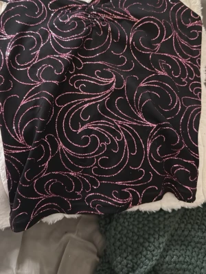 Svart tubtopp med rosa glittermönster Orsay - Snygg svart tubtopp från Orsay med glittrigt rosa mönster i swirl-design. Perfekt för dig som vill sticka ut med lite extra glitter. Toppen är axelbandslös och har en croppad passform.