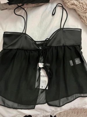 Svart transparent linnetopp Zara (prislapp kvar) - Säljer en svart linnetopp från Zara med tunna axelband och transparent, flowy material. Toppen har en söt snörning framtill och en glansig kant upptill som ger en extra touch. Perfekt för dig som vill ha en trendig och luftig topp till sommaren.🖤