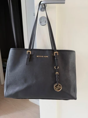 Svart handväska från Michael Kors - Stilren svart handväska från Michael Kors i ett tåligt material med guldfärgade detaljer och logotyp framtill. Väskan har två långa axelband och en rund MK-berlock som hänger på framsidan. Perfekt storlek för att få plats med allt du behöver till jobbet eller skolan!! Min dator får plats och det är ett fack i mitten som delar upp sidorna, perfekt. Använd många gånger vilket syns på banden, men den har inga skador i övrigt. Väldigt bra skick!💓Nypris är 4200 kr!!