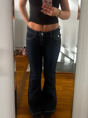 Mörkblå bootcut jeans med vita sömmar - Säljer ett par mörkblå bootcut jeans med vita kontrastsömmar och låg midja. Jeansen har fickor fram och bak med knappdetaljer på bakfickorna. Storlek 170 och endast använda ett fåtal gånger🩷