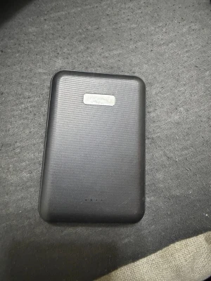 Svart trådlös powerbank med MagSafe - Säljer en svart powerbank med MagSafe-funktion för trådlös laddning. Den har en slät, rektangulär form och är tillverkad i plast. Perfekt för att ladda mobilen på språng utan sladdar.