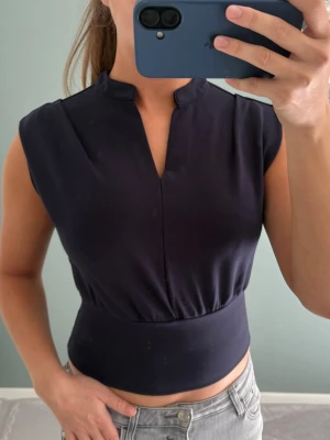 Svart croppad topp med ståkrage - Snygg croppad topp med liten ståkrage och v-ringad öppning framtill. Toppen är ärmlös och har midjeresår som ger en fin passport 