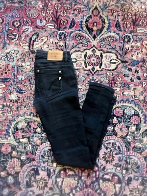 Svart jeans från Dondup - Snygga svarta jeans från Dondup med klassisk femficksdesign och detaljer i metall. Jeansen har en skinny/slim passform och är tillverkade i ett mjukt denimtyg med läderpatch bak i midjan. Jeansen har även ett litet hål vid grenen men inget man ser när man bär de. Perfekta för en trendig och avslappnad look.