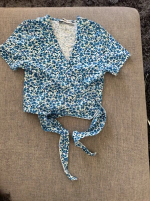Blommig omlottopp från AB Sister - Söt omlottopp från AB Sister i vitt och blått med blommigt mönster. Toppen har korta ärmar, v-ringning och knytband i midjan. Perfekt för dig som gillar en feminin och trendig stil.