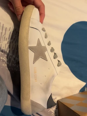 Vita sneakers med grå stjärna - Säljer ett par vita sneakers med grå mockadetaljer och en grå stjärna på sidan. Skorna har grå snören och beige sula. Märket GGDB/STAR syns på sidan och plösen. Perfekta för dig som gillar stilrena och trendiga sneakers.