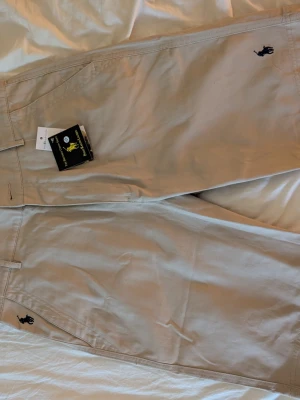RL Beige shorts med broderad logga - Snygga beige shorts med en mörkblå broderad logga på sidan. Klassisk design med raka ben och diskreta sömmar. Perfekta för varma dagar och enkel att matcha med olika stilar.