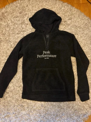 Svart Peak Performance hoodie  - Mysig svart hoodie från Peak Performance med broderad logga på bröstet. Tillverkad i fluffigt teddy-material, har huva, halv dragkedja och stor magficka framtill. Perfekt för dig som gillar en avslappnad och sportig stil.