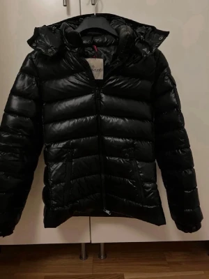 Svart jacka Moncler - Jackan har huva, dragkedja framtill och två fickor med dragkedja. Insidan har Monclers klassiska tidnings porträtt. I storlek 2. PRIS KAN DISKUTERAS