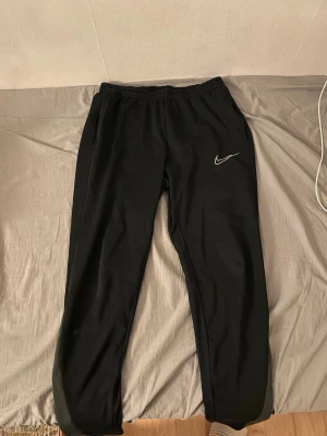 Svarta mjukisbyxor från Nike - Svarta mjukisbyxor från Nike med klassisk vit logga på vänster lår. Byxorna har resår i midjan och smalare benavslut. Perfekta för chill eller träning. Materialet känns mjukt och stretchigt.