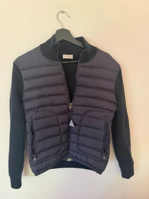 Mörkblå dunväst från Moncler - Snygg mörkblå cardigan från Moncler med quiltad framsida och två dragkedjeförsedda fickor. Västen har ribbad krage och mudd vid ärmslut och nederkant. Perfekt för lager på lager och ger en nice look.