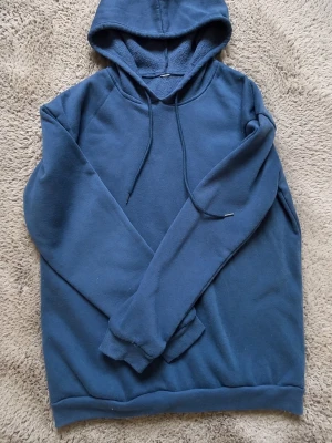 Blå hoodie med huva och dragsko - Mysig blå hoodie med huva och dragsko. Tillverkad i mjukt material med borstad insida för extra komfort. Klassisk modell med långa ärmar och ribbade muddar vid ärmslut och nederkant. Perfekt för en avslappnad och chill stil.
