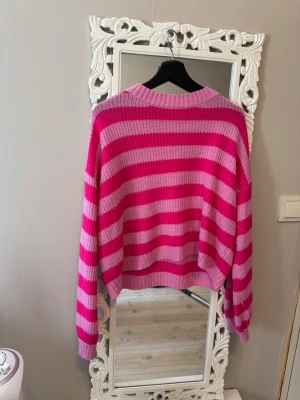 Rosa och ceriserandig stickad tröja - Säljer en stickad tröja med breda ränder i rosa och cerise. Tröjan har avslappnad passform, rund halsringning och långa ärmar. Perfekt för dig som gillar färg och vill ha en mysig, trendig look.