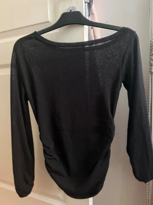 Svart långärmad topp med rynk - En svart långärmad topp med rund halsringning och rynkade detaljer på ena sidan. Tunt, lätt material som ger en snygg siluett. Perfekt att styla med jeans eller kjol för en trendig look.