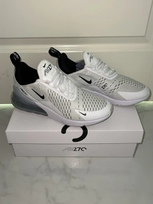 Nike Air Max 270 vita sneakers - Nike Air Max 270 sneakers i vitt med svarta detaljer och genomskinlig Air Max-häl. Ovandelen är i mesh för bra andningsförmåga och skon har en sportig, modern look med tydliga loggor på plös och sidor. Perfekt för dig som gillar streetwear och stilrena sneakers.
