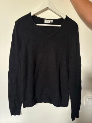Svart V-ringad stickad tröja Vila - Klassisk svart stickad tröja från Vila med V-ringning och långa ärmar. Tröjan har ribbade muddar vid ärmslut och nederkant, vilket ger en snygg och enkel look. Perfekt basplagg till garderoben.