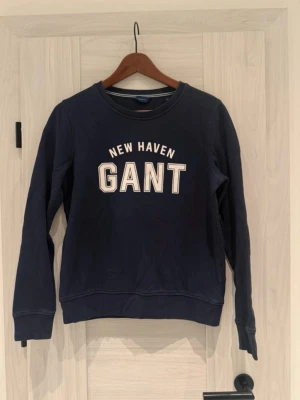 Mörkblå GANT "New Haven" Sweatshirt - Säljer en jättefin marinblå sweatshirt från GANT med det klassiska ”New Haven”-trycket på bröstet. Tröjan har en stilren och tidlös passform. Perfekt för både vardag och mer preppy tillfällen! Endast sparsamt använd, så den är i mycket gott skick. • Märke: GANT • Storlek: S • Färg: Marinblå / Navy • Skick: Mycket gott skick • Detaljer: Logga framtill, mjuk ruggad insida, ribbade muddar. • Skickar inom 48 timmar! 🚚 • Rök och djurfritt hem. 🚭🐶
