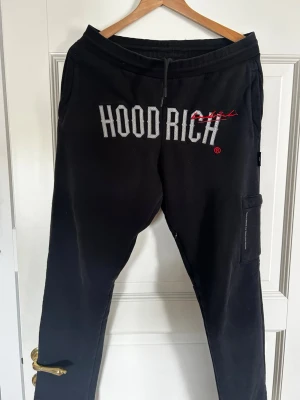 Svarta mjukisbyxor från Hoodrich - Svarta mjukisbyxor från Hoodrich med stor vit logga framtill och röd detalj. Byxorna har resår i midjan med dragsko och en ficka med dragkedja på ena benet. Perfekta för en avslappnad streetwear-look. Pris kan diskuteras!