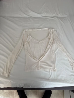 Vit långärmad kofta från H&M - Säljer en tunn, vit långärmad kofta från H&M. Koftan har en djup V-ringning och spetsdetaljer vid halsen. Materialet är mjukt och stretchigt, perfekt att slänga över ett linne eller topp. Stängs med en knapp framtill.