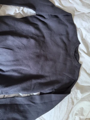 Mörkgrå enkel stickad tröja - En basic mörkgrå stickad tröja med rund halsringning och långa ärmar. Tröjan har en klassisk passform och ribbade muddar vid halsen. Perfekt att matcha med jeans eller kjol för en clean look.