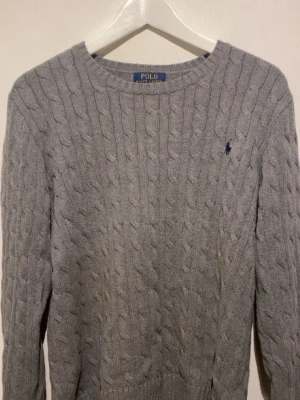 Grå kabelstickad tröja Polo Ralph Lauren - Kabelstickad grå tröja från Polo Ralph Lauren med rund halsringning och klassisk broderad logga på bröstet. Långa ärmar och ribbade muddar ger en snygg och tidlös look. Perfekt för lager-på-lager-stil. Priset är inte hugget i sten, släng iväg ett dm om du har några frågor 😁